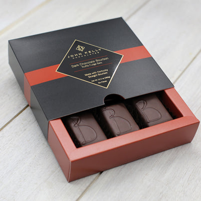 A square, red box sits partially slide out from a black box. The black box has a red stripe going horizontally in the middle with a black John Kelly Chocolates label. The red box holds three, rectangular pieces of chocolate with the letter B on all of them.
