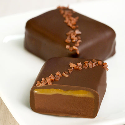 Two squares of chocolate sit on a white plate. They both have red-ish salt on them. One is cut revealing the caramel and chocolate inside.
