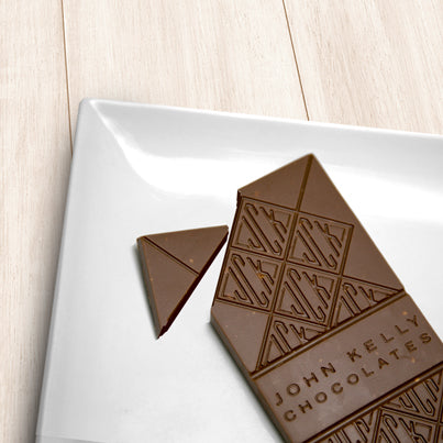 A milk chocolate solid bar with the top left corner cut off from it. The bar has a John Kelly Chocolates logo on it.