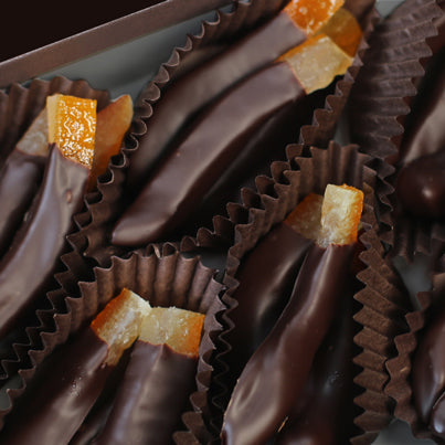A close up of the chocolate dipped orange peels in the box. They are sitting in groups of two in brown, paper cups. The orange peels are mostly covered in chocolate, leaving less than one fourth of the peel visible.