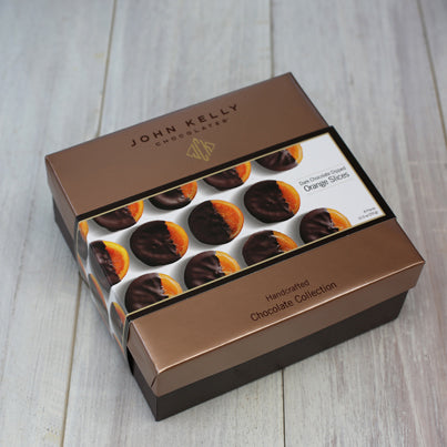 A box of the chocolate covered orange slices. The box is brown in color and sits closed. A white sleeve holds the box closed. The sleeve has pictures of the orange slices neatly laid out.