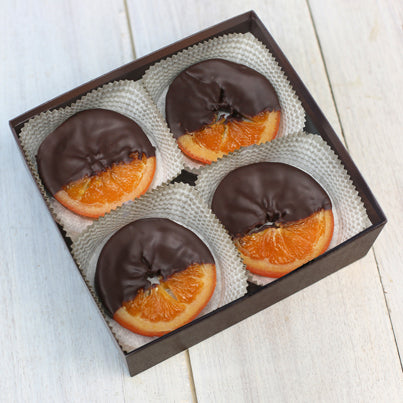 A open with four chocolate covered orange slices in it. The orange slices are sitting in white, paper cups. The orange slices are covered in chocolate, leaving one fourth of the orange slice uncovered.