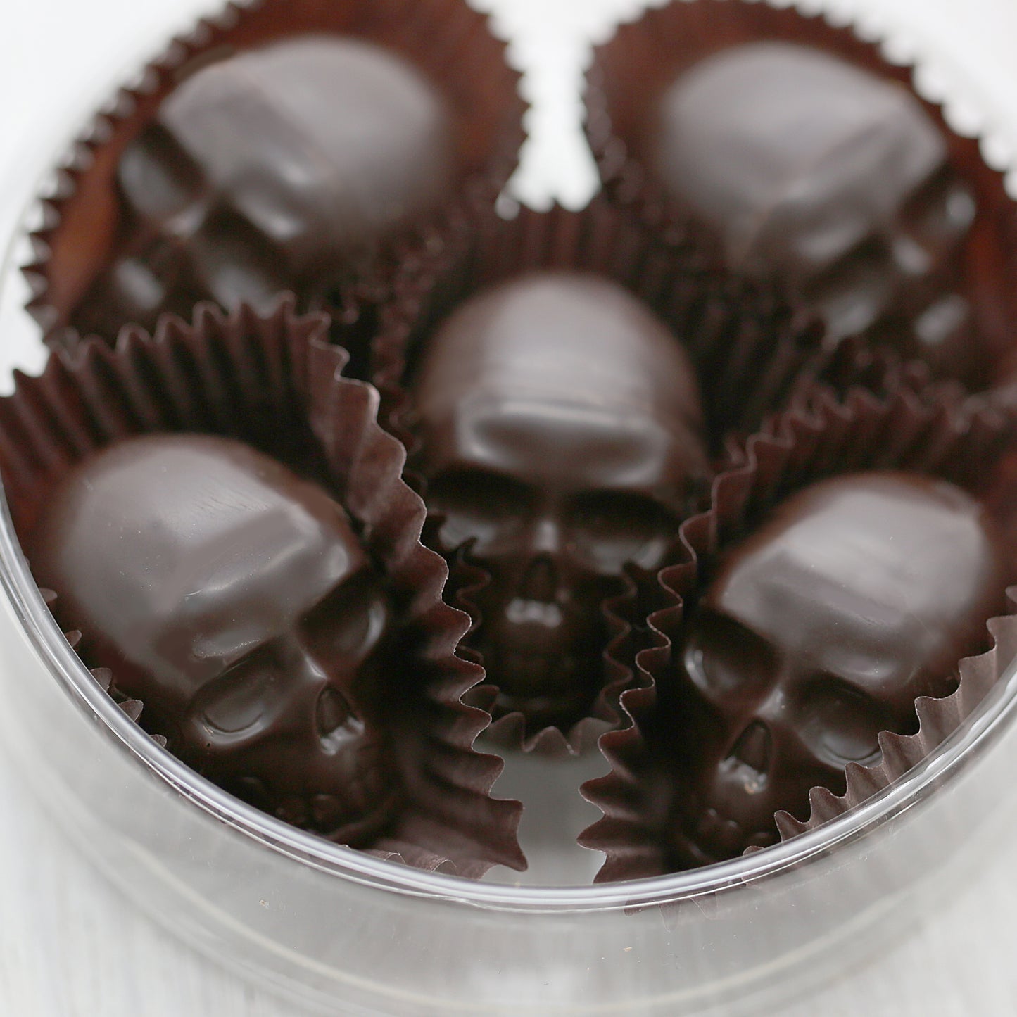 Five small dark chocolates skulls with brown paper cups around them. They are placed in a clear, round, plastic container