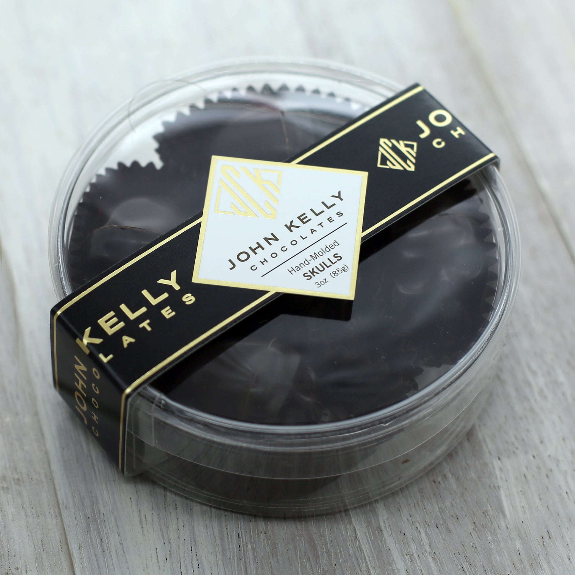A closed five piece small dark chocolate skull container. The container is clear, plastic, and round and is closed with a black John Kelly Chocolates logo