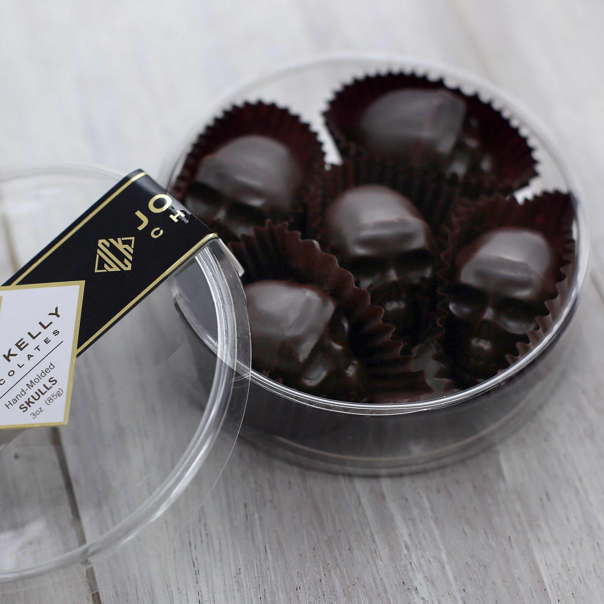 Five small dark chocolate skulls in a round, plastic container. The lid of the container is leaned against the container and has a black John Kelly Chocolates logo