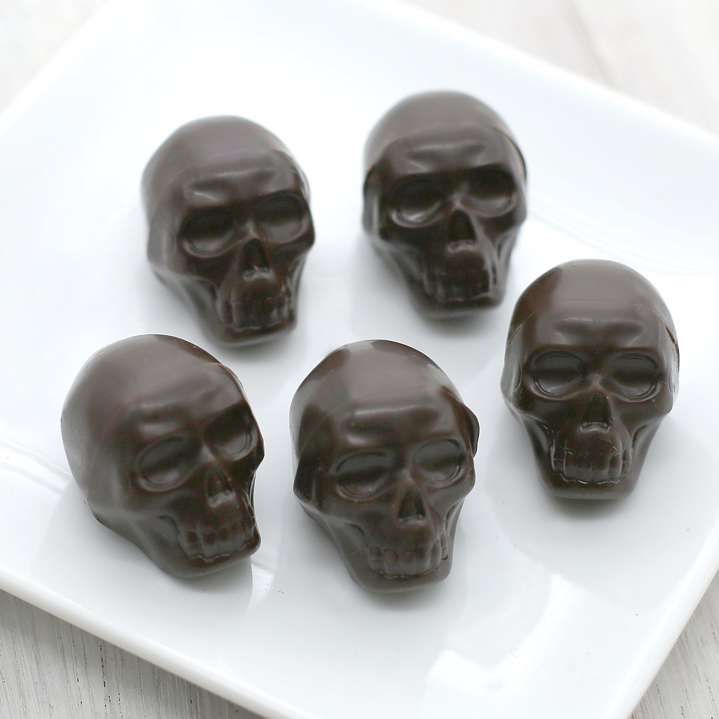 Five dark chocolate skulls sit on a white, square plate.