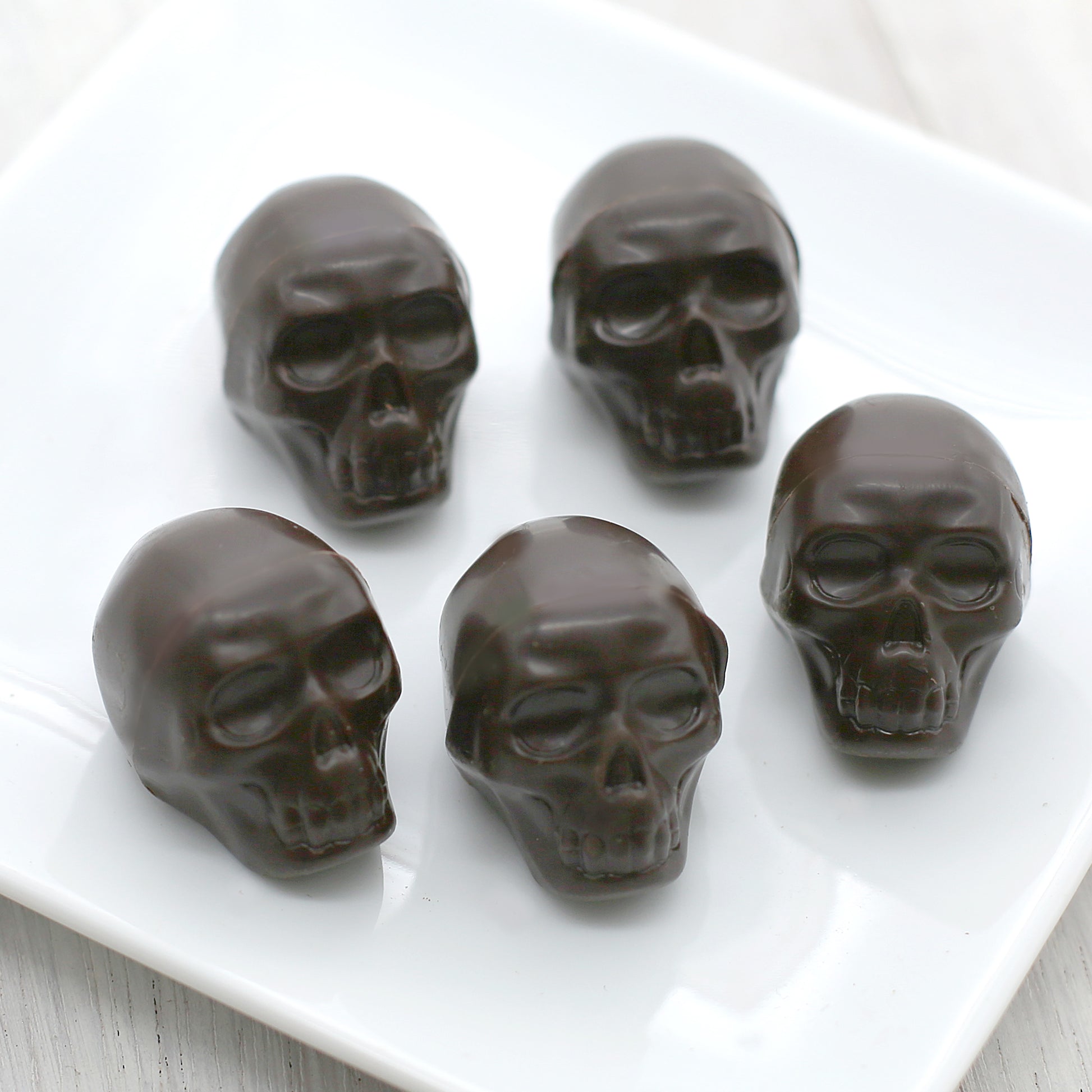 Five dark chocolate skulls sit on a white, square plate.