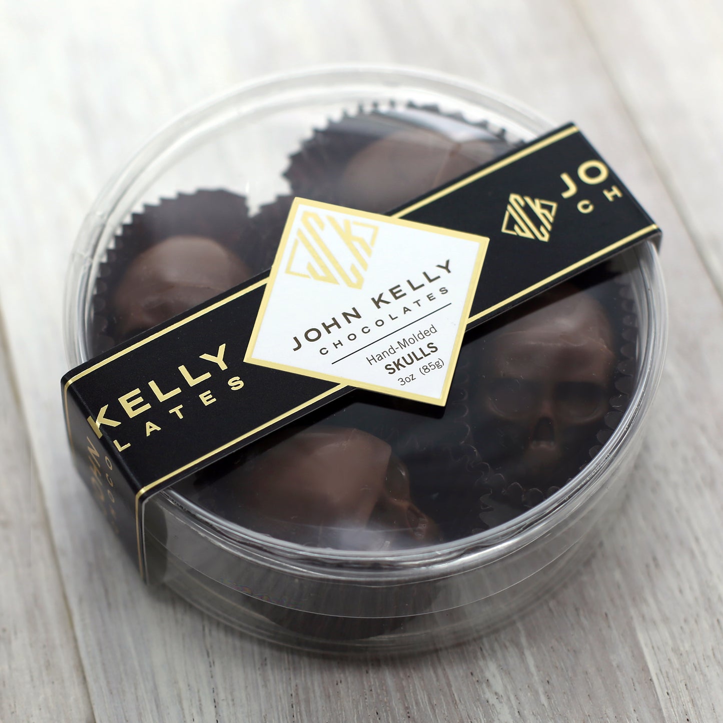 A closed container of the five piece milk chocolate skulls container. The container is clear, round, plastic, and has a black John Kelly Chocolates logo on it.