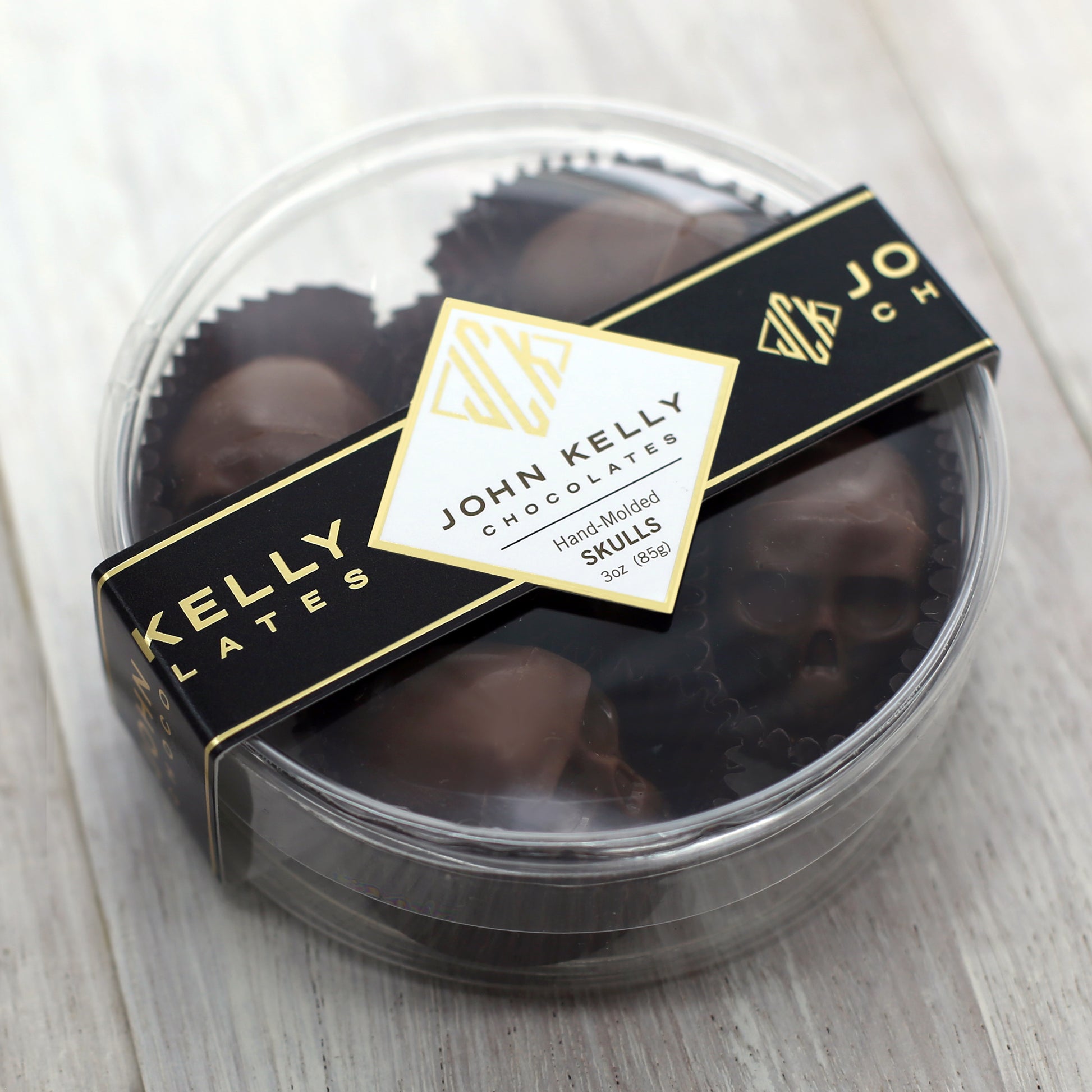 A closed container of the five piece milk chocolate skulls container. The container is clear, round, plastic, and has a black John Kelly Chocolates logo on it.