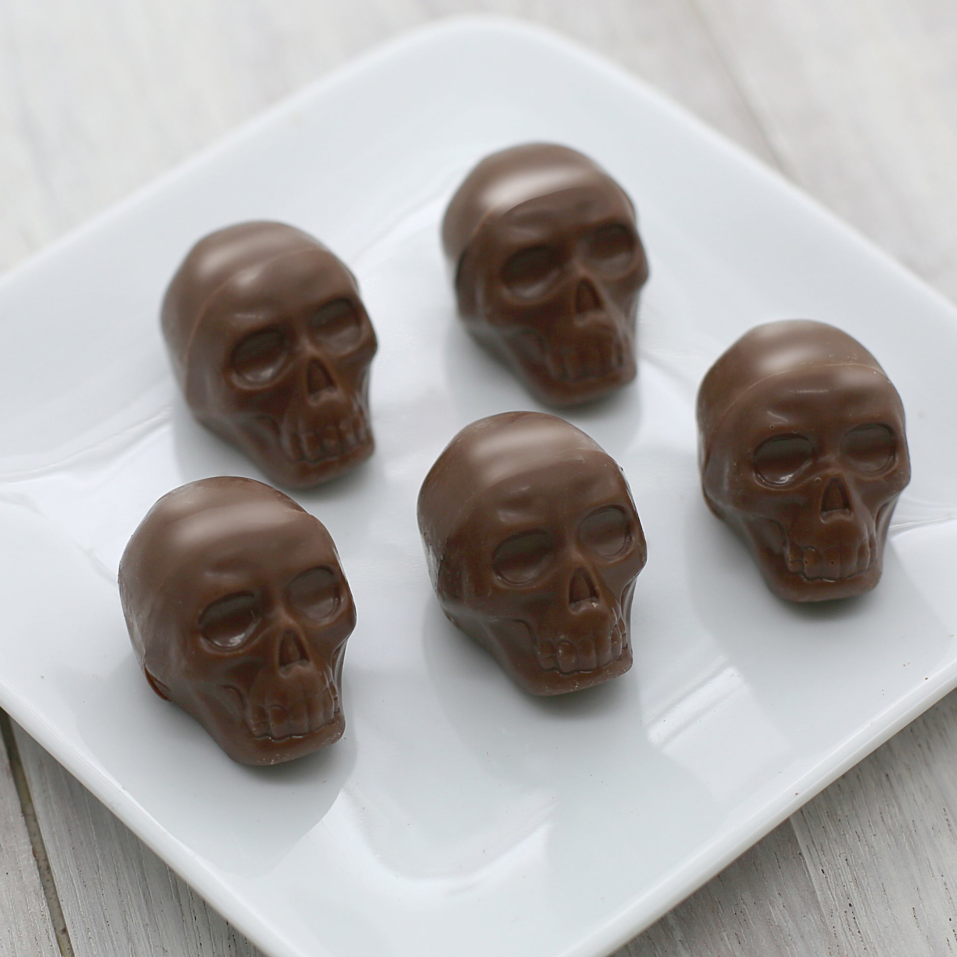 Five milk chocolate skulls sit on a white, square plate.